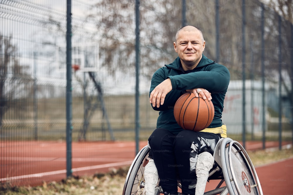 Portrait of sportsman in wheelchair holding basketball on outdoor court and looking at camera.