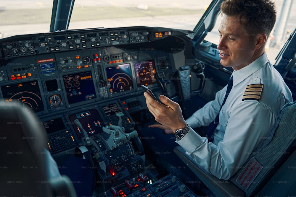 Giovane pilota maschio sorridente e concentrato con un cellulare in mano seduto nella cabina di pilotaggio