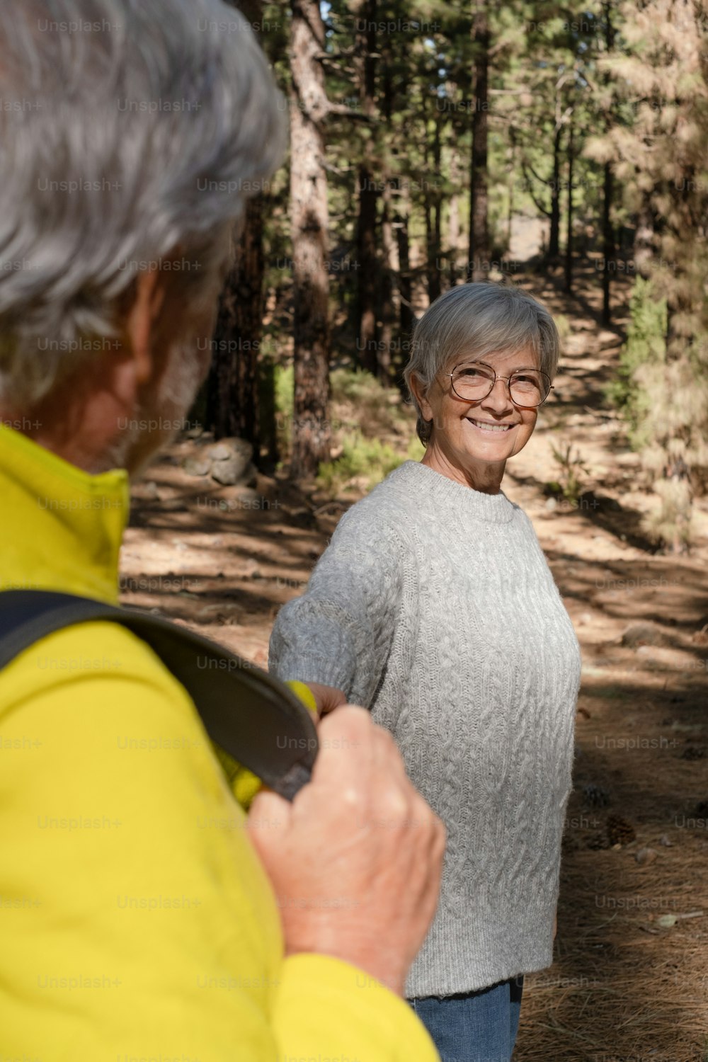 手をつないで森の中を歩く夫を見つめる魅力的な年配の女性。白人のアクティブな年配の白髪の人々は、休暇の自由と歩道での自然ハイキングを楽しんでいます
