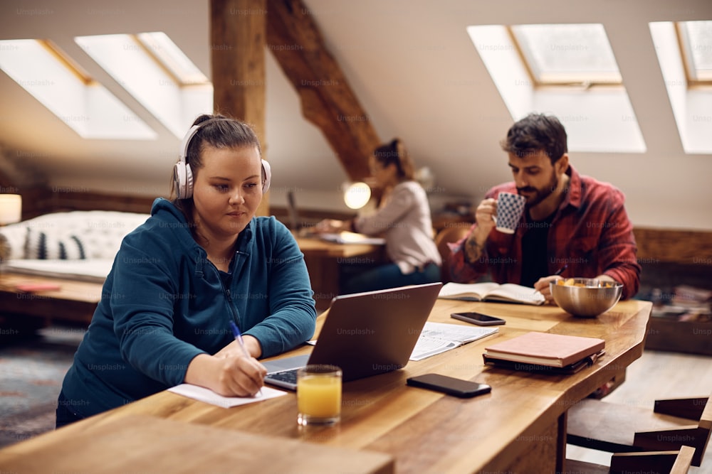 Estudiante con auriculares tomando notas mientras aprende en línea en una computadora en casa. Sus amigos estudian en el fondo.