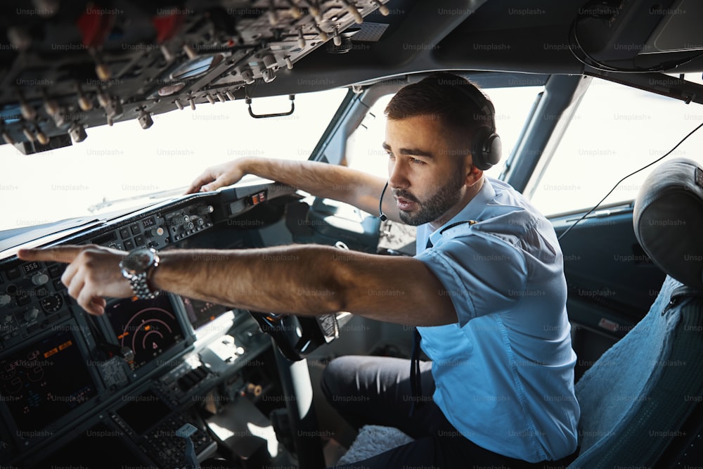 Un homme caucasien sérieux dans un cockpit montrant quelque chose derrière la vitre avant le début du vol