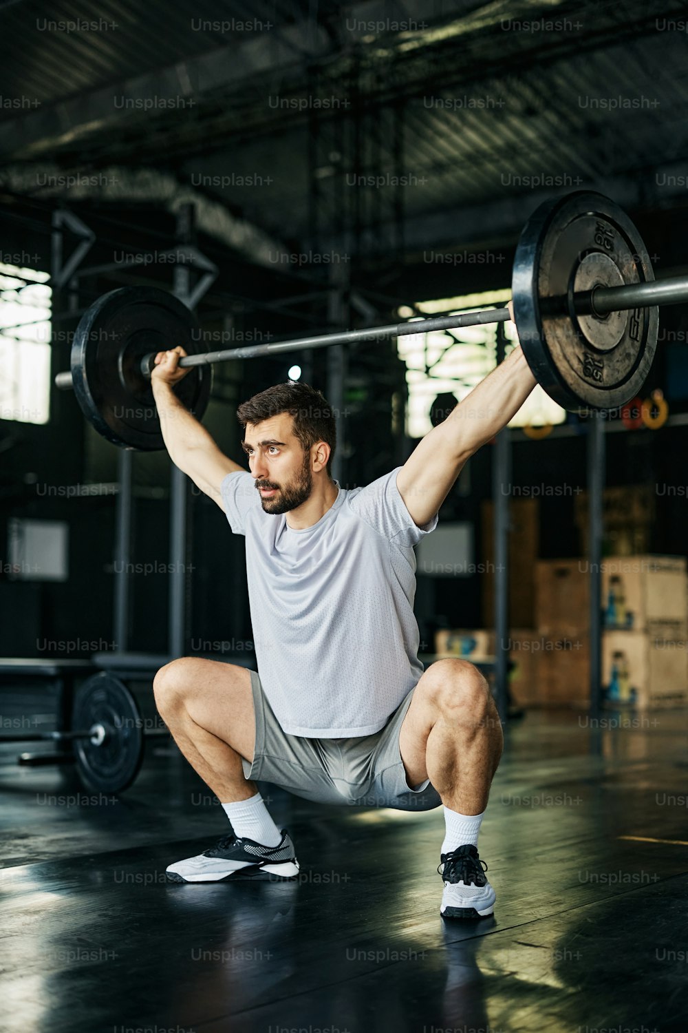 Male athlete holding barbell above his head while doing squats during cross training in a gym.
