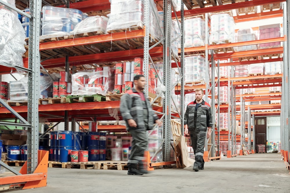 Two young intercultural male workers in uniform moving along huge shelves with packed goods inside large factory or warehouse