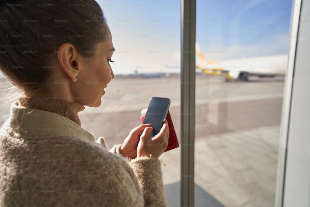 Woman pressing on screen of her mobile phone while holding flight documents before window on airport runway