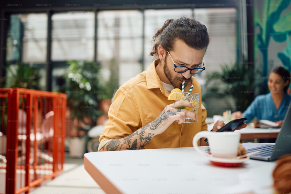Männlicher Unternehmer trinkt Cocktail, während er in einem Café sein Handy benutzt.