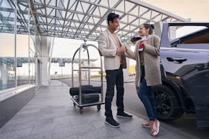 Adult male in eyeglasses looking at female, standing near car and holding passport and boarding pass in hand