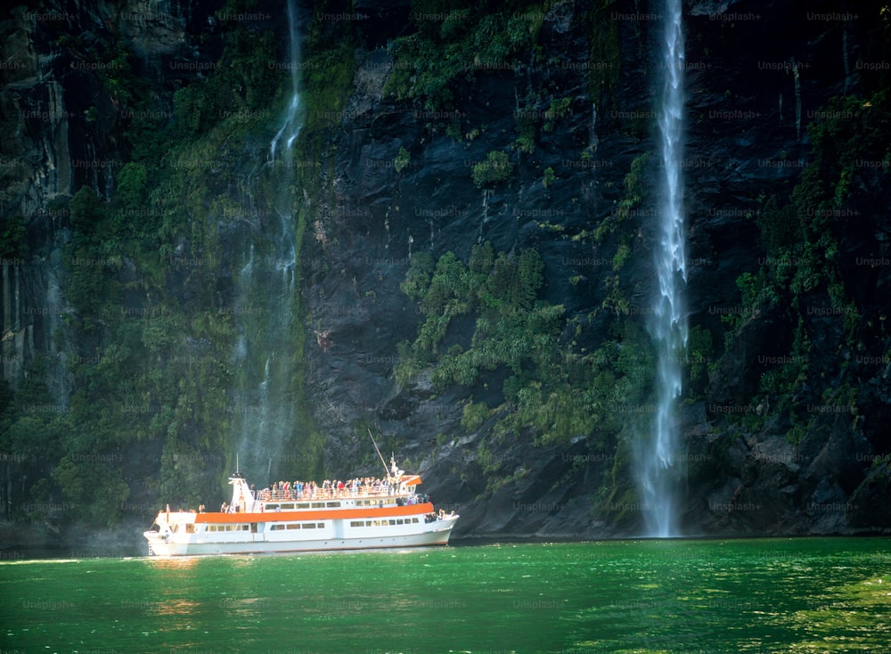 Sightseeing boat carrying tourist people approaches great waterfall in Milford Sound. Beautiful scenic cruise through Fiordland National Park in South Island of New Zealand.