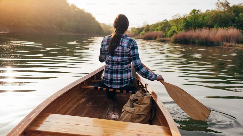 Rear view of hipster girl paddling the canoe on the sunset lake.