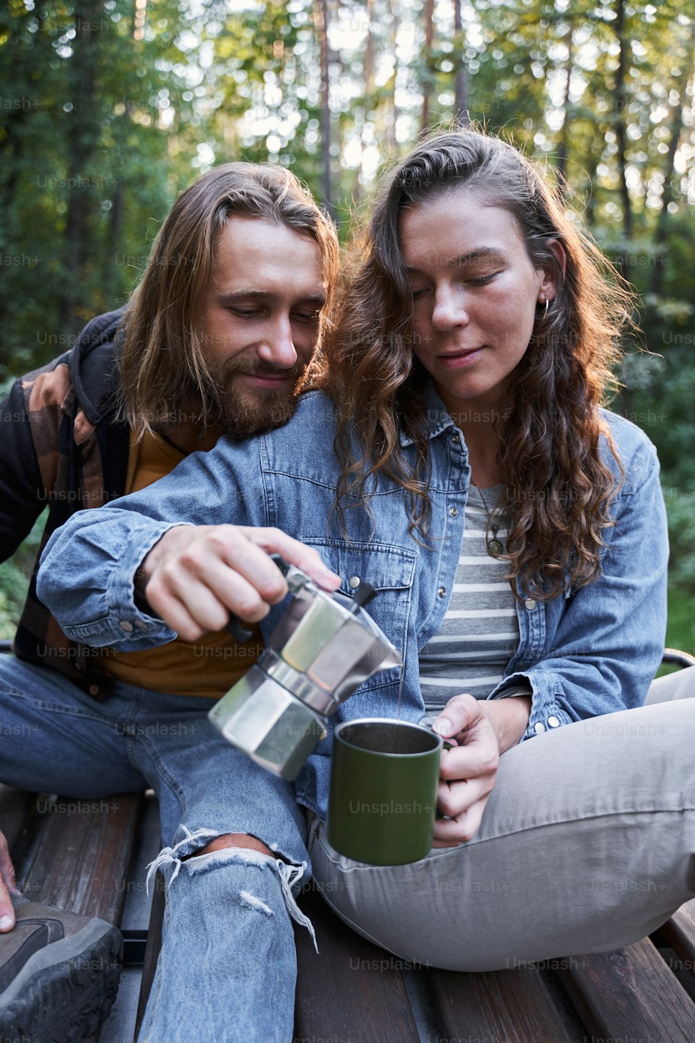 Bearded man looking at the cup while his girlfriend pouring coffee in his cup at campsite. Couple camping in nature and having coffee