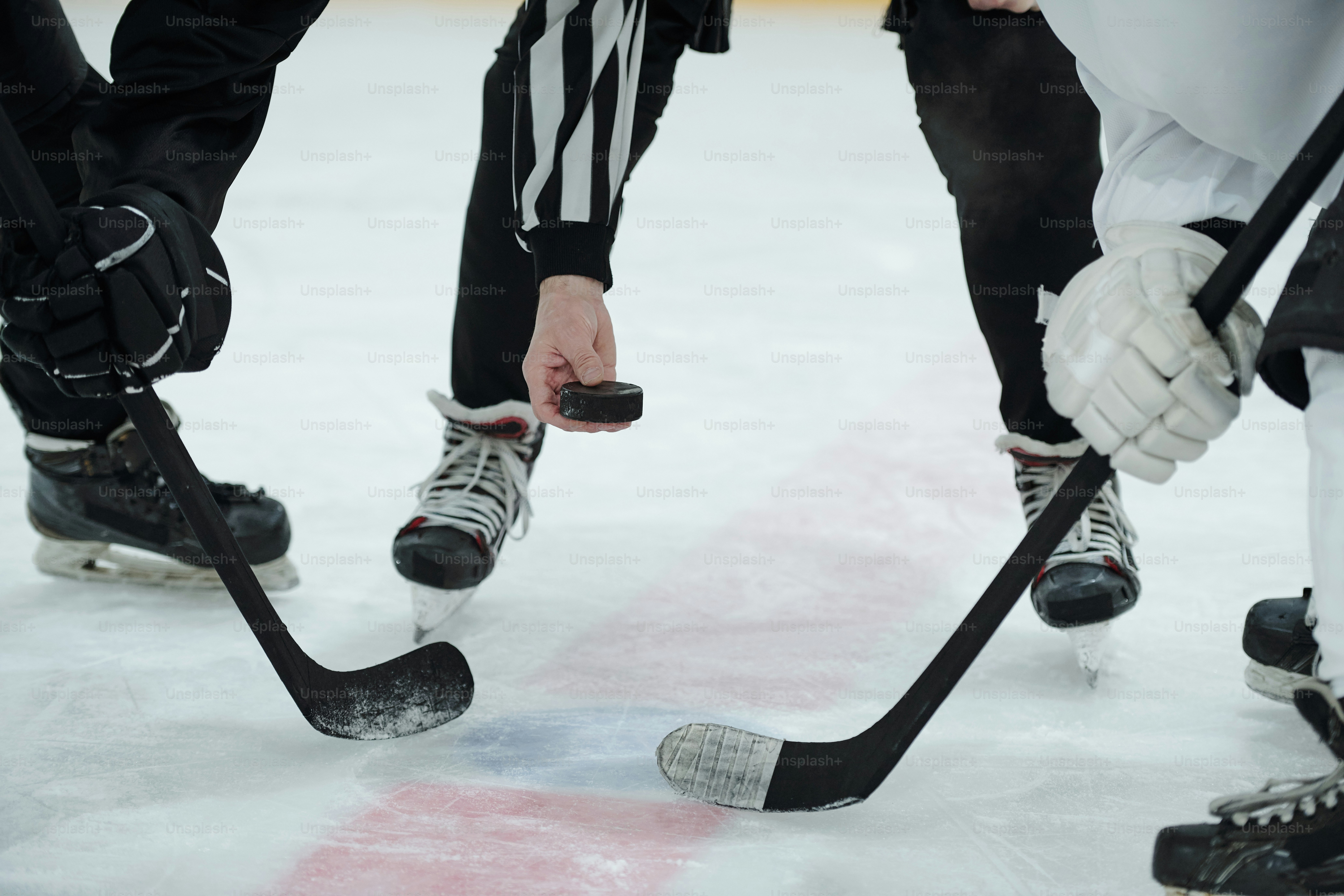 Hand of referee holding puck over ice rink with two hockey players with sticks standing around him and waiting for moment to shoot it
