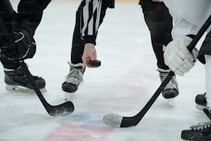 Hand des Schiedsrichters, der den Puck über die Eisbahn hält, während zwei Eishockeyspieler mit Stöcken um ihn herum stehen und auf einen Moment warten, um ihn zu schießen