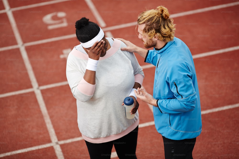Professional sport instructor encouraging and supporting anxious over-size female after workout on stadium before marathon