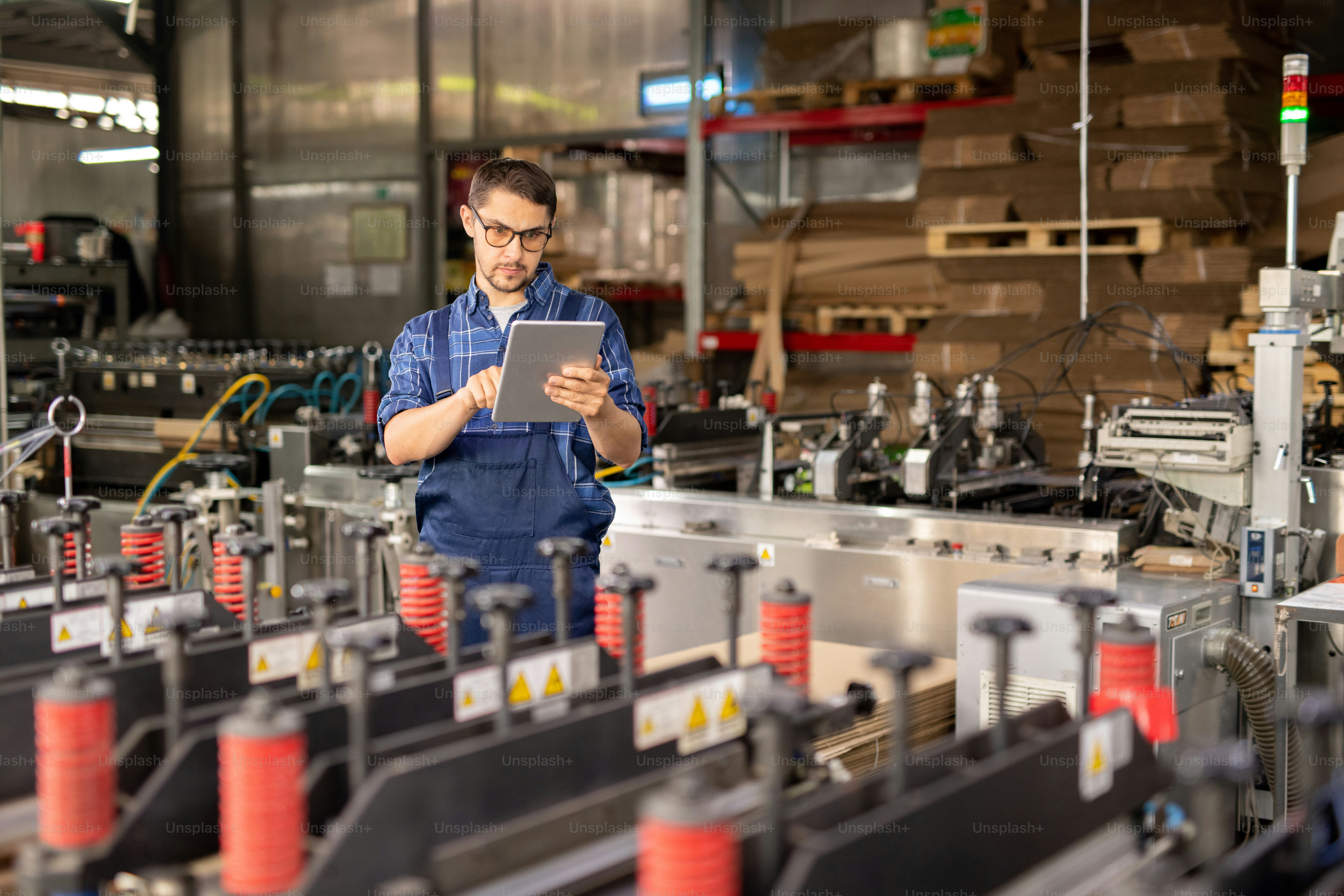 Engenheiro contemporâneo da planta industrial usando touchpad enquanto está de pé ao lado de grande máquina industrial na oficina
