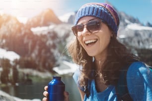 Portrait of handsome smiling hiking woman standing in stunning mountain wilderness with bottle near snowy lake