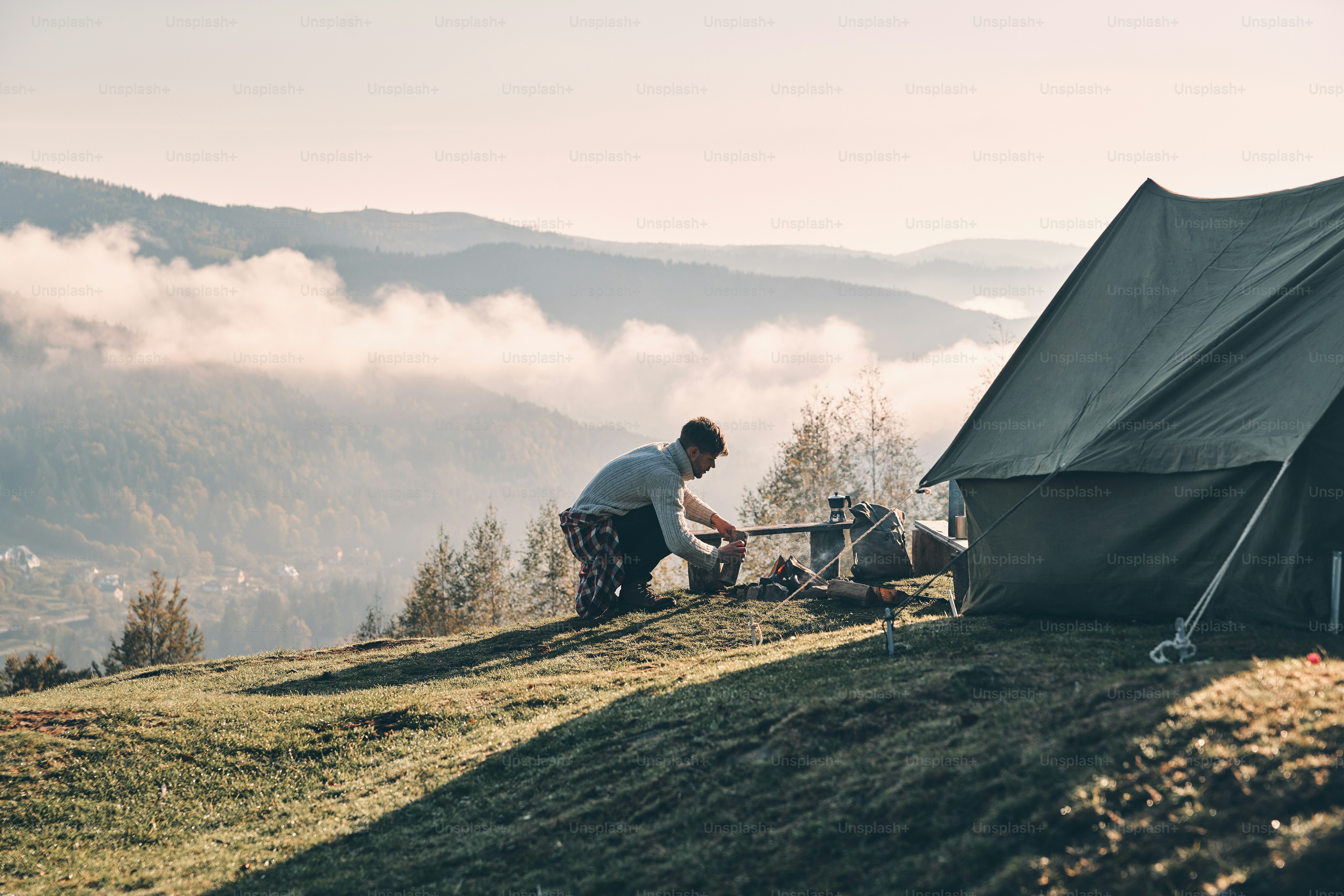 Young man making a campfire while sitting near the tent in the ...