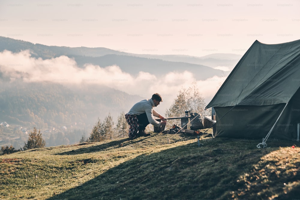 Young man making a campfire while sitting near the tent in the mountains