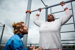 Young trainer encouraging plump female to try harder in doing pull-ups on bar