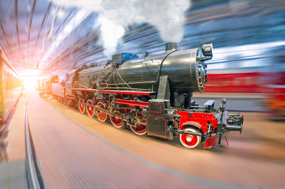 An old retro locomotive train railroad with steam and smoke clubs arrives at the passenger covered station under the roof