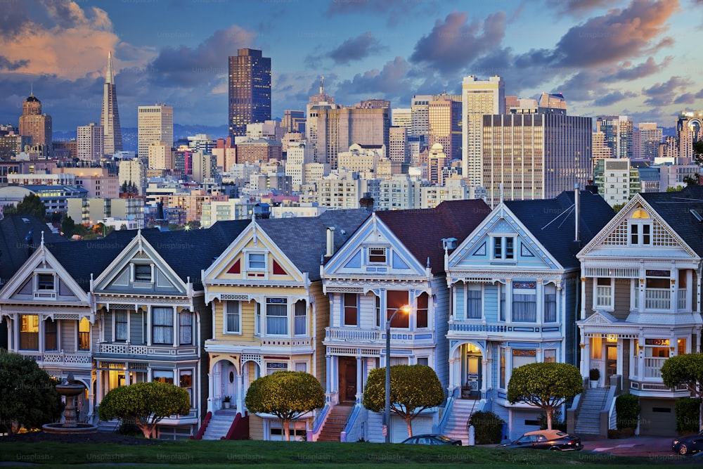 Famous Painted Ladies of San Francisco, California sit glowing amid the backdrop of a sunset and skyscrapers.