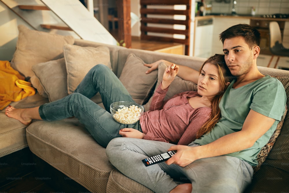 Junges Paar isst Popcorn, während es sich auf dem Sofa entspannt und im Wohnzimmer fernsieht.