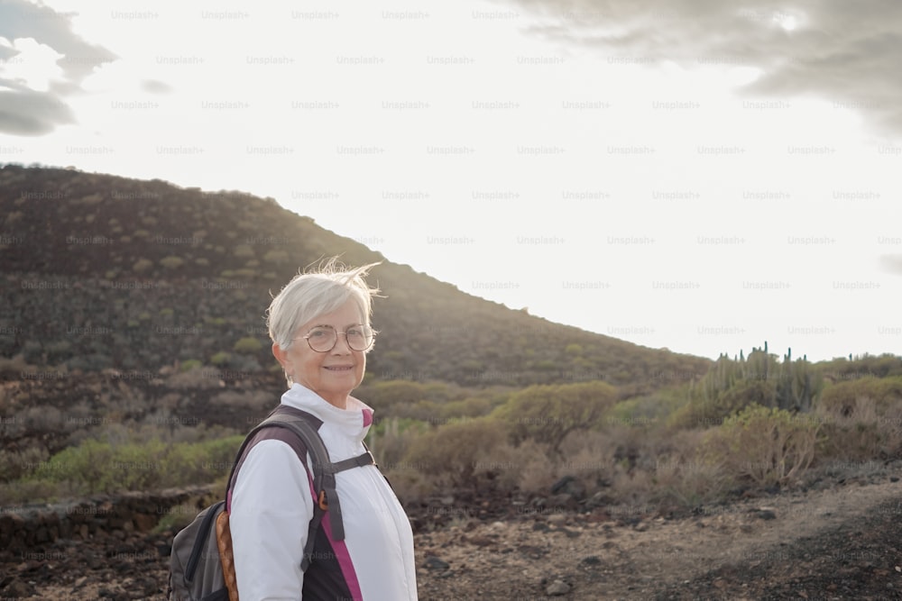 Donna anziana attraente sorridente dai capelli grigi che cammina in escursione all'aperto con zaino sulle spalle. Donna anziana spensierata che si gode la libertà e uno stile di vita sano