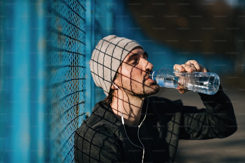 Tired athletic man drinking water with eyes closed while taking a break from exercising outdoors.