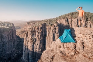 L'escursionista dell'uomo o l'alpinista estremo si trova vicino alla tenda da campeggio su un punto panoramico della scogliera nel profondo canyon di Tazi in Turchia. Scopri le meraviglie della natura e il concetto di avventura pericolosa