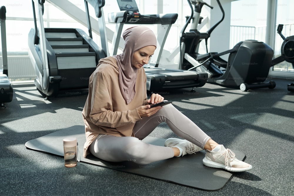 Horizontal shot of young adult Muslim woman wearing hijab sitting on mat in gym watching something on her smartphone during break time