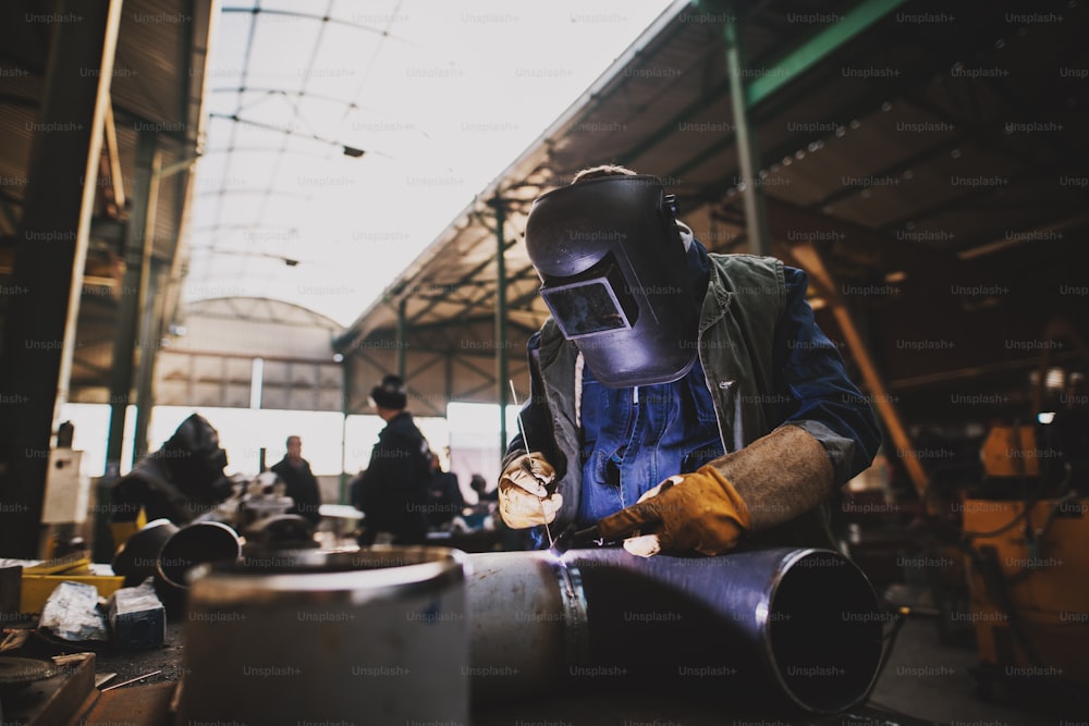 Soldador con uniforme protector y máscara soldando tubería metálica en la mesa industrial mientras saltan chispas.