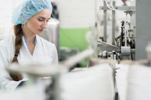 Happy female employee wearing protective headwear and white lab coat while working as a manufacturing engineer in a contemporary cosmetics factory