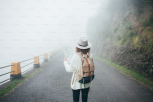 Fille voyageuse bouclée portant un sac à dos et un chapeau debout sur la route dans le brouillard, à la recherche d’un sentier et à l’aide d’une carte.