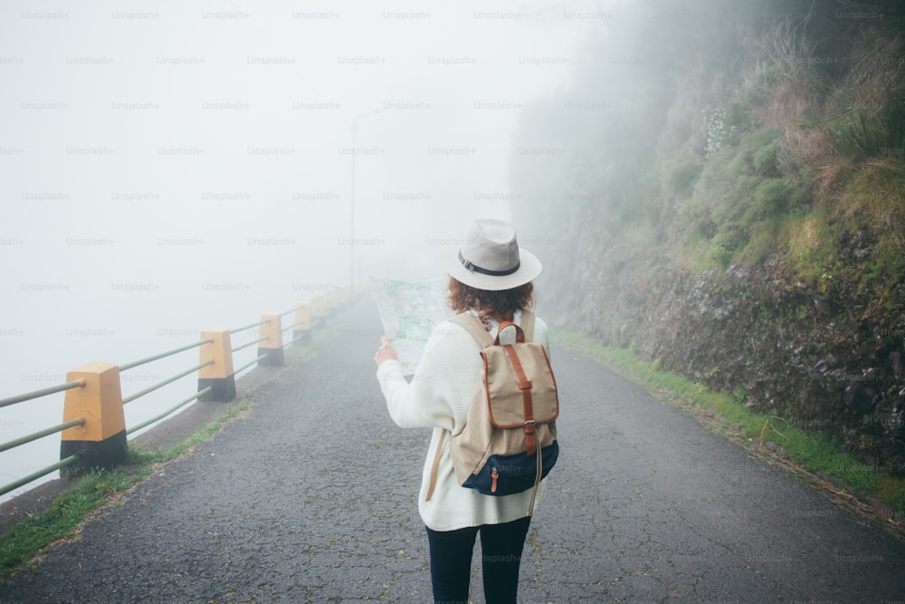 Menina viajante encaracolada usando mochila e chapéu em pé na estrada em neblina, procurando trilha e usando mapa.
