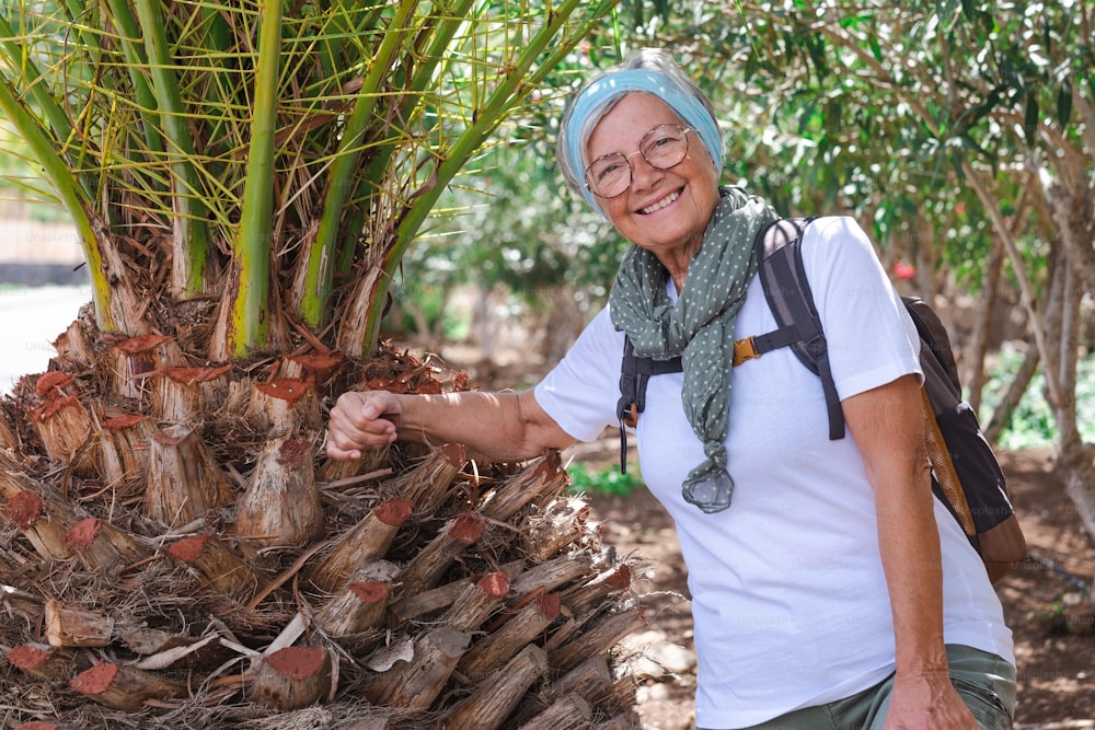Happy active senior woman with backpack in outdoors excursion enjoying healthy lifestyle and sunny day.  Elderly attractive female in tropical woods smiles looking at camera