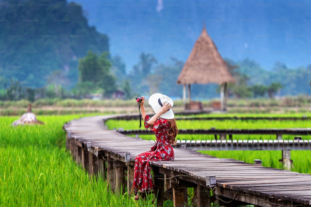 Young woman sitting on wooden path and take a photo by camera with green rice field in Vang Vieng, Laos.