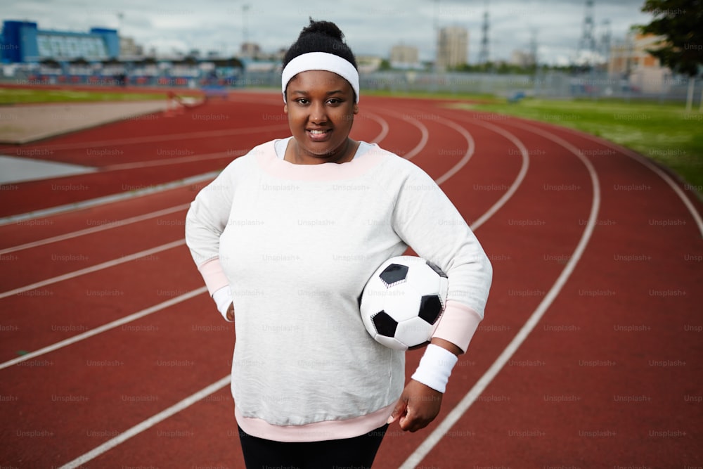 Retrato de mujer regordeta positiva posando con pelota de fútbol en el estadio de atletismo
