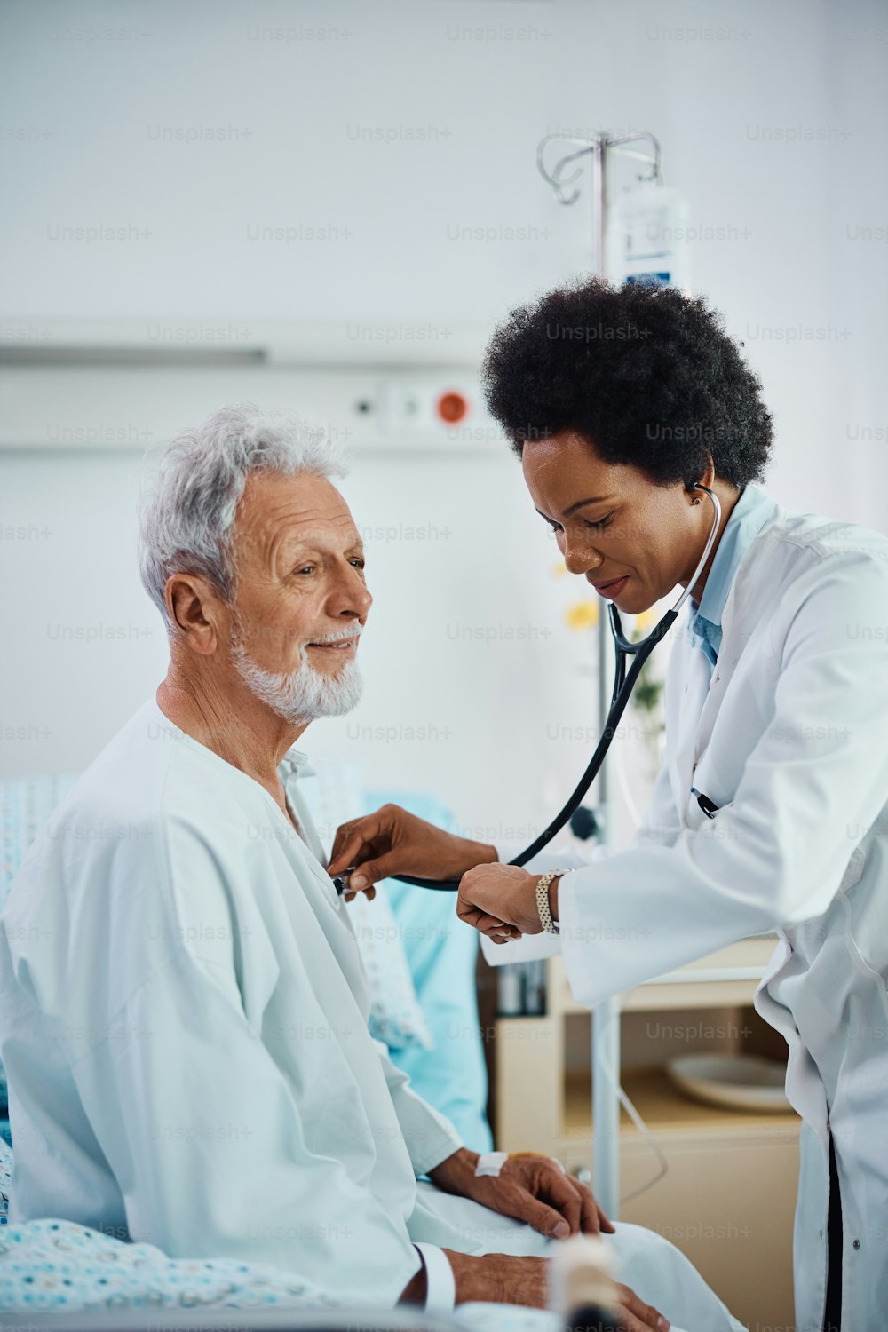 Cardiologo afroamericano sorridente che usa lo stetoscopio mentre visita il suo paziente anziano nel reparto ospedaliero.