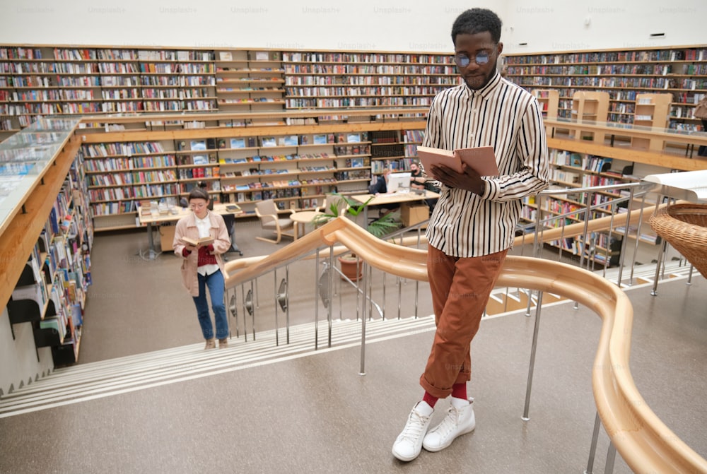 African young man in eyeglasses reading a book while standing in the big library