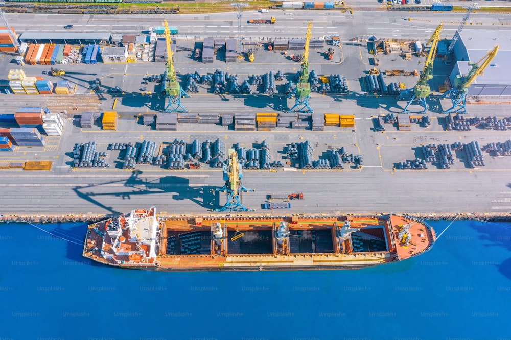 Aerial top view huge cargo ship moored at the pier at the port, loading goods, metal in aluminum rolls, concrete and other solid raw materials.