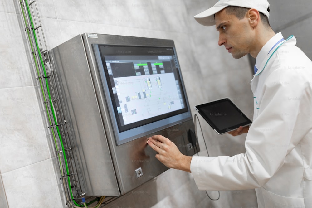Technologist in a white coat makes the necessary entries in the tablet is at the factory. The man selects the desired parameters on the digital touch screen on the device in the shop. Worker operating pasteurizer using the control panel