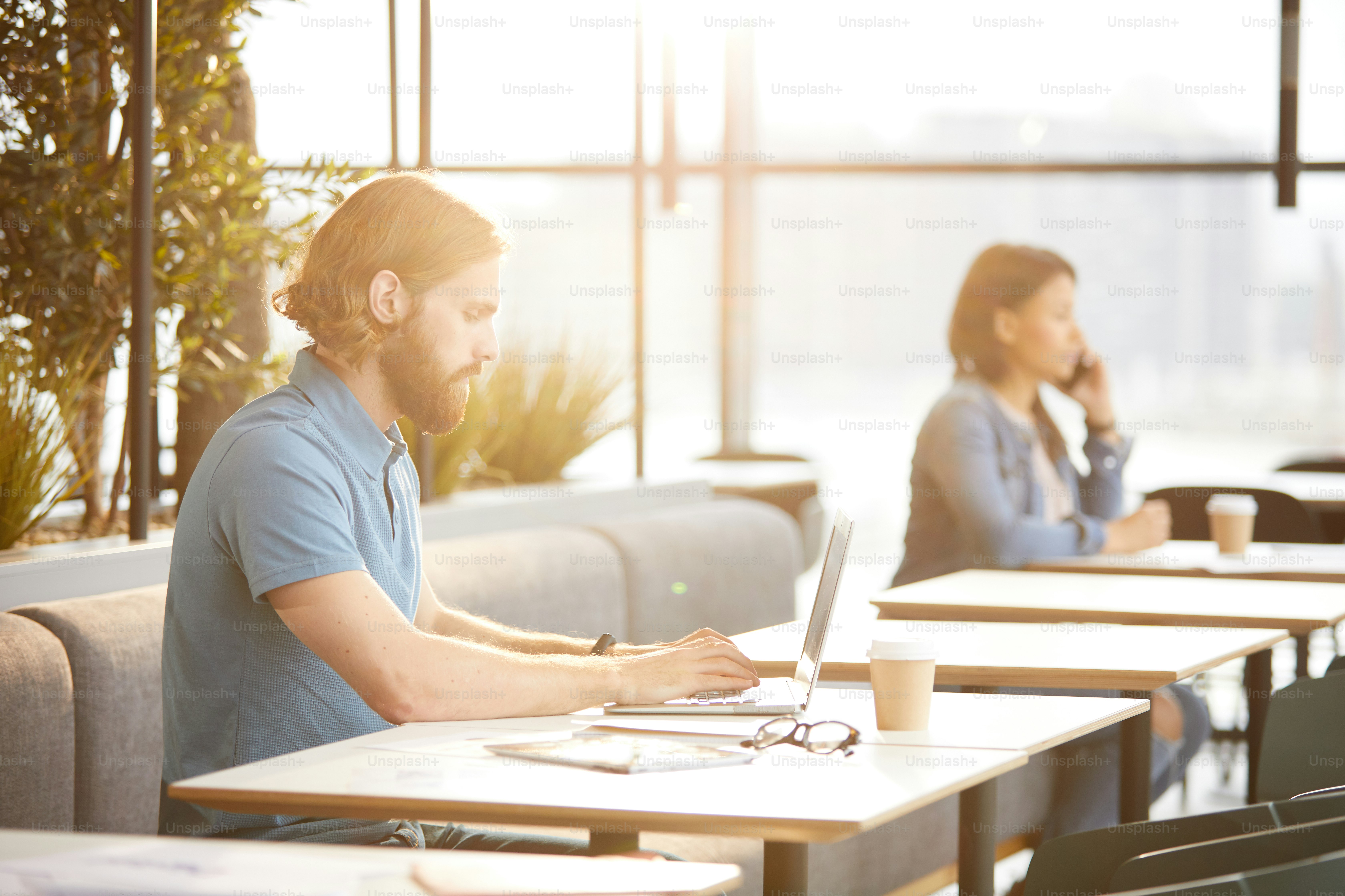 Young bearded businessman in casual clothing working on laptop computer while sitting at the table in cafe with businesswoman in the background