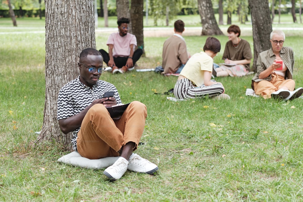 African young businessman sitting on the grass outdoors and using his mobile phone at work with other people in the background