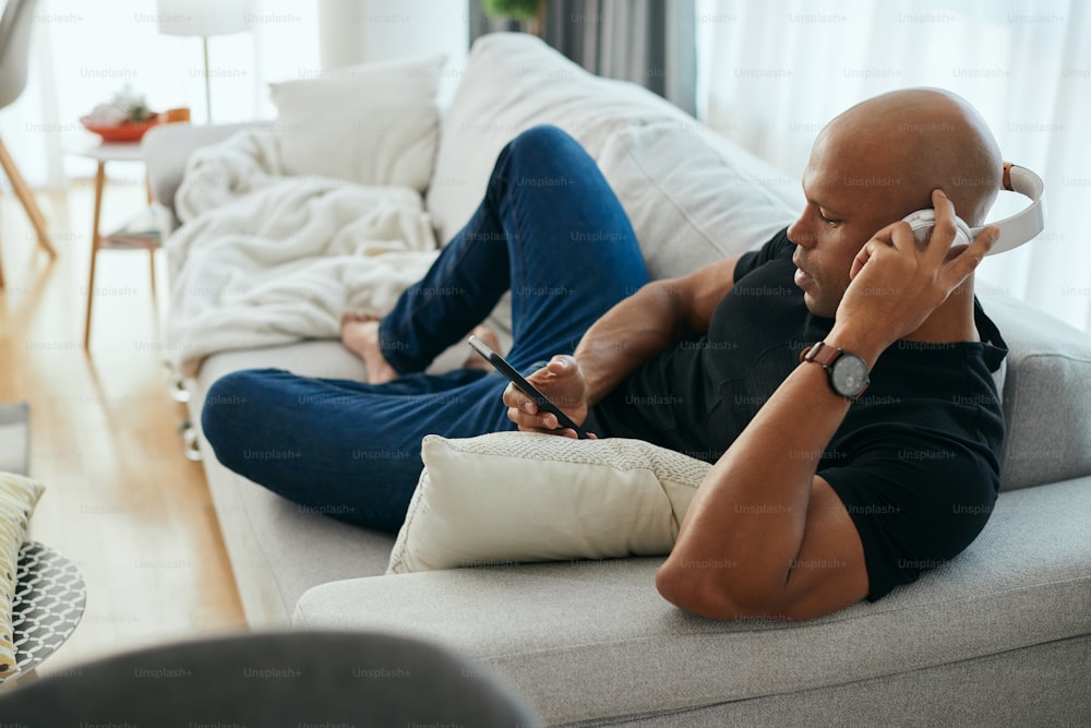 Young African American man wearing headphones while using smart phone and relaxing on the sofa at home.