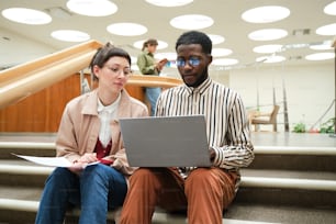 African man using laptop and studying together with woman while they sitting on stairs in the library