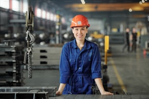 Portrait of smiling young Asian metalworking specialist in blue overalls standing at desk in industrial shop