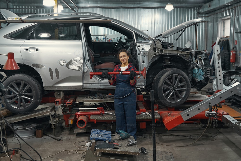 Full length of woman automobile technician holding metal tool while standing near lifted car in repair service station