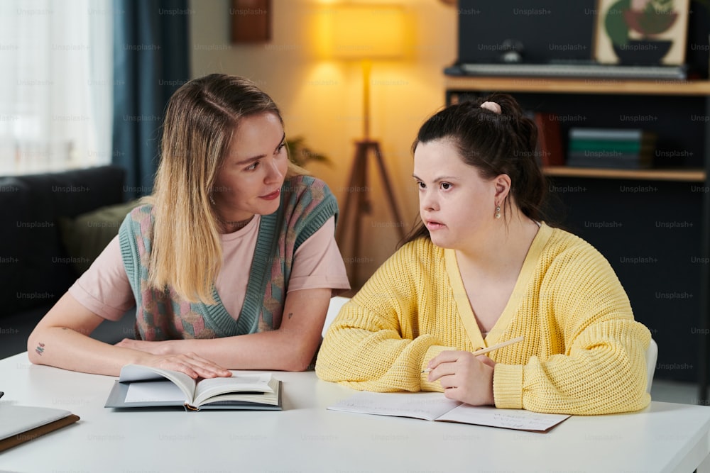 Young Caucasian woman working as teacher sitting at table next to female student with Down syndrome attentively listening to her during individual class