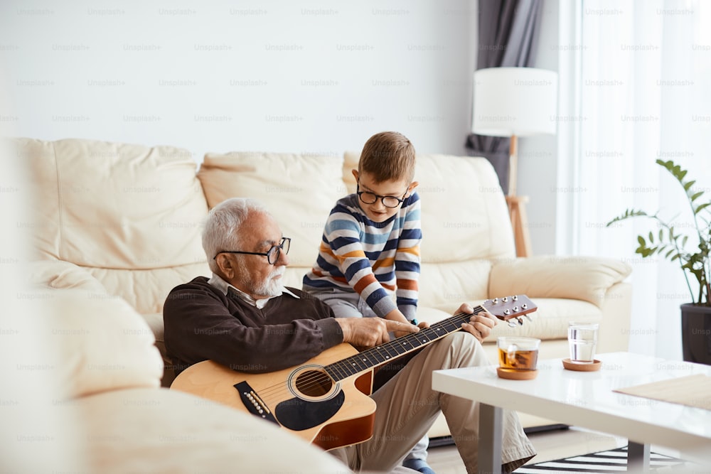 Älterer Mann genießt mit seinem Enkel und bringt ihm bei, zu Hause akustische Gitarre zu spielen.