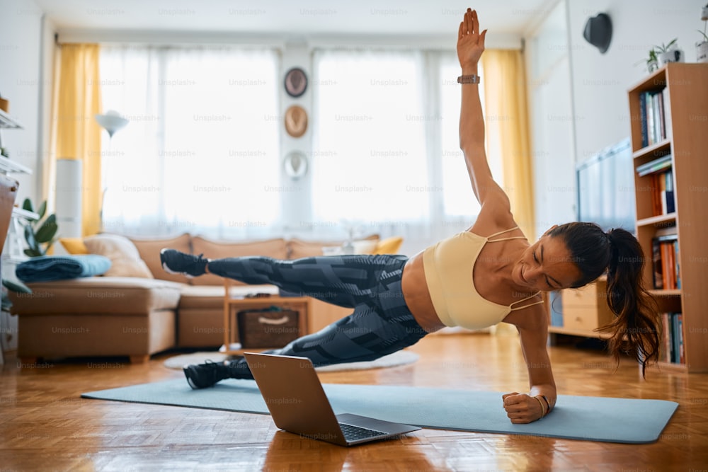 Instrutor de fitness segurando aula de exercícios via laptop de casa e fazendo prancha lateral com perna e braço levantados durante o treino.