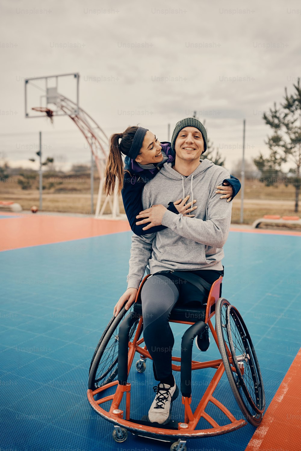 バスケットボールコートで車椅子を使用している障害のある男性の友人を抱きしめる幸せな運動女性。