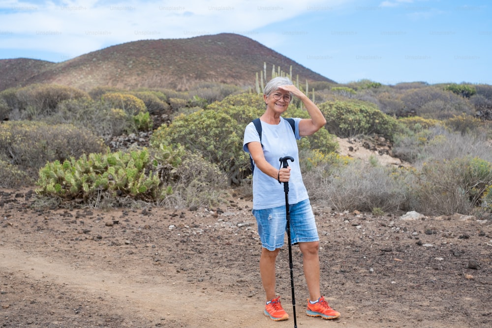 Donna anziana sorridente in escursione all'aperto che trasporta uno zaino e un bastone da passeggio. Vecchia donna in forma durante un'escursione che si gode la libertà dell'avventura e una vacanza sana